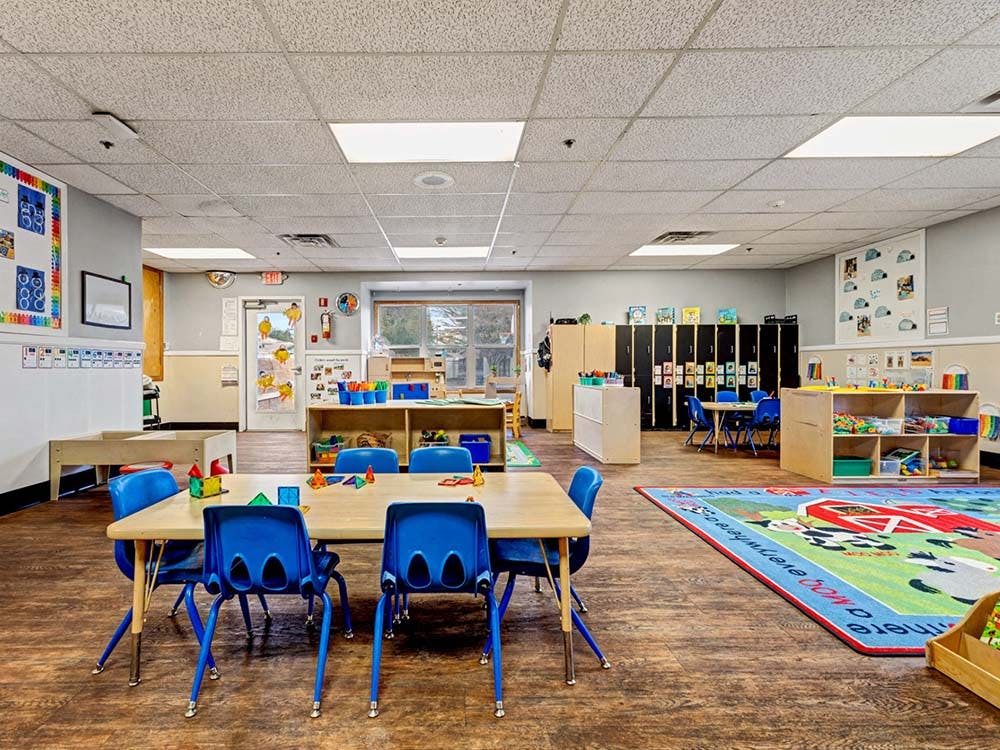 Classroom with tables, blue chairs, shelves with toys, and a colorful rug