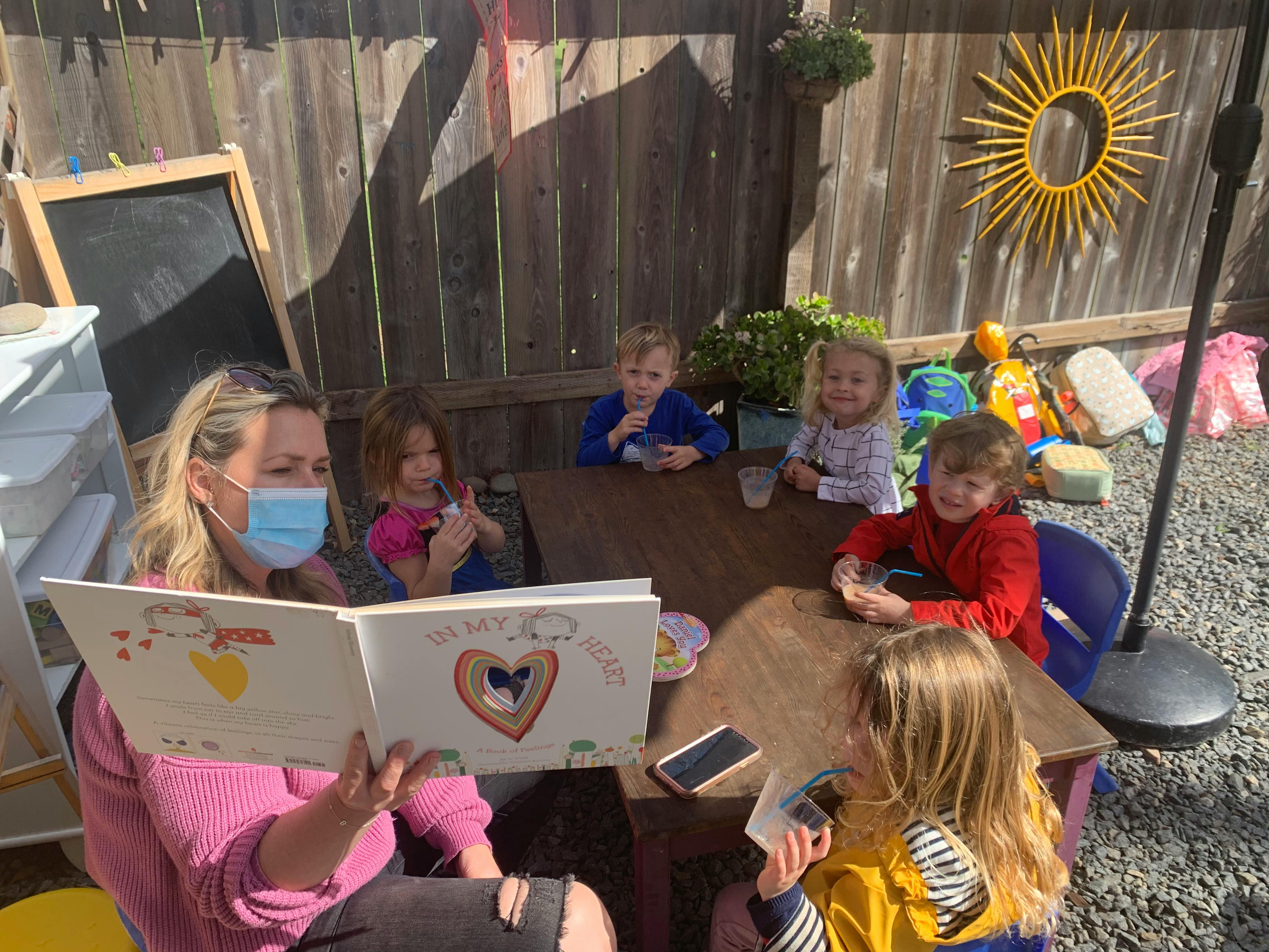 Person in mask reading a book to children at a table outdoors
