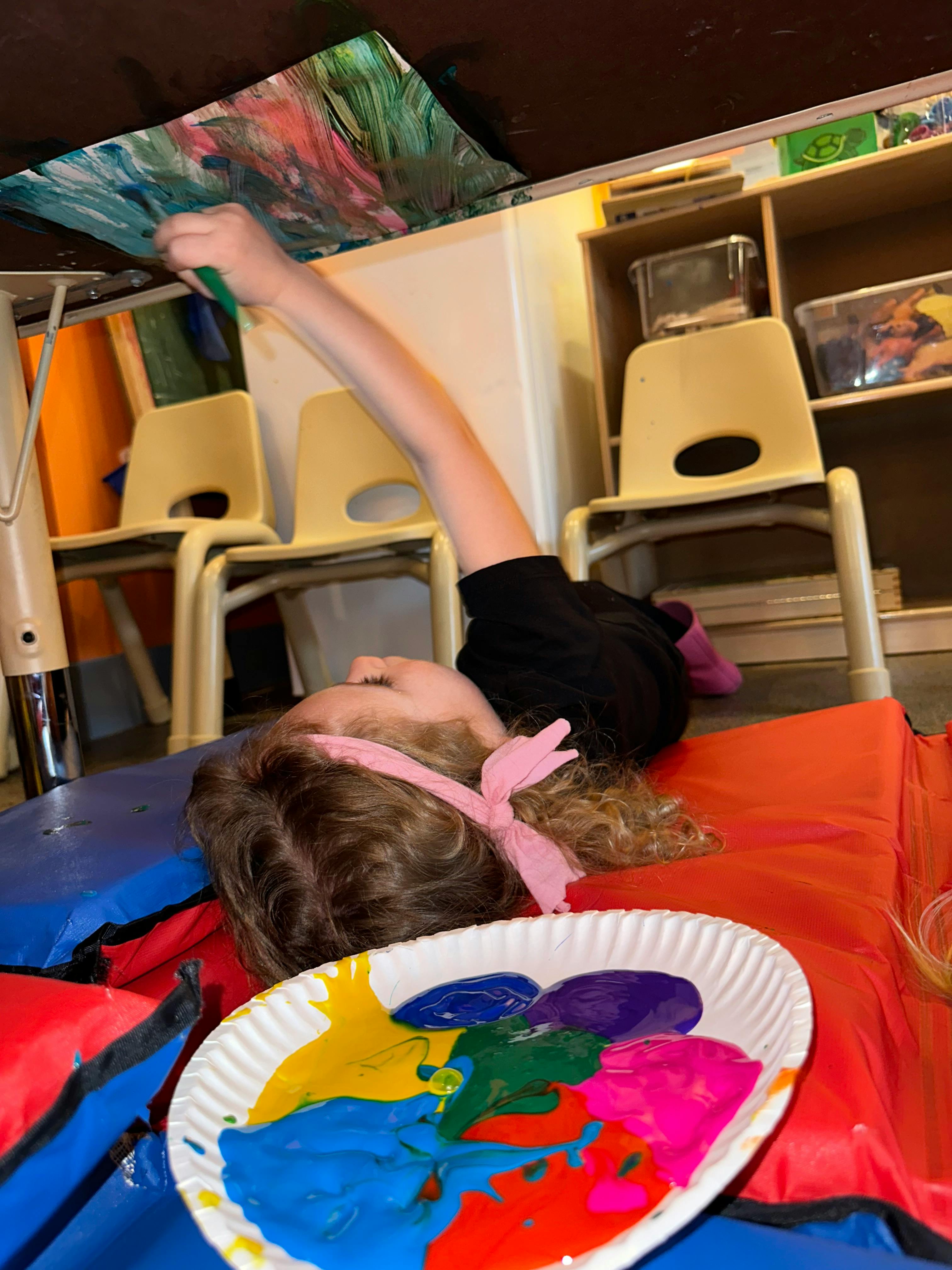 Child lying on back painting under a table