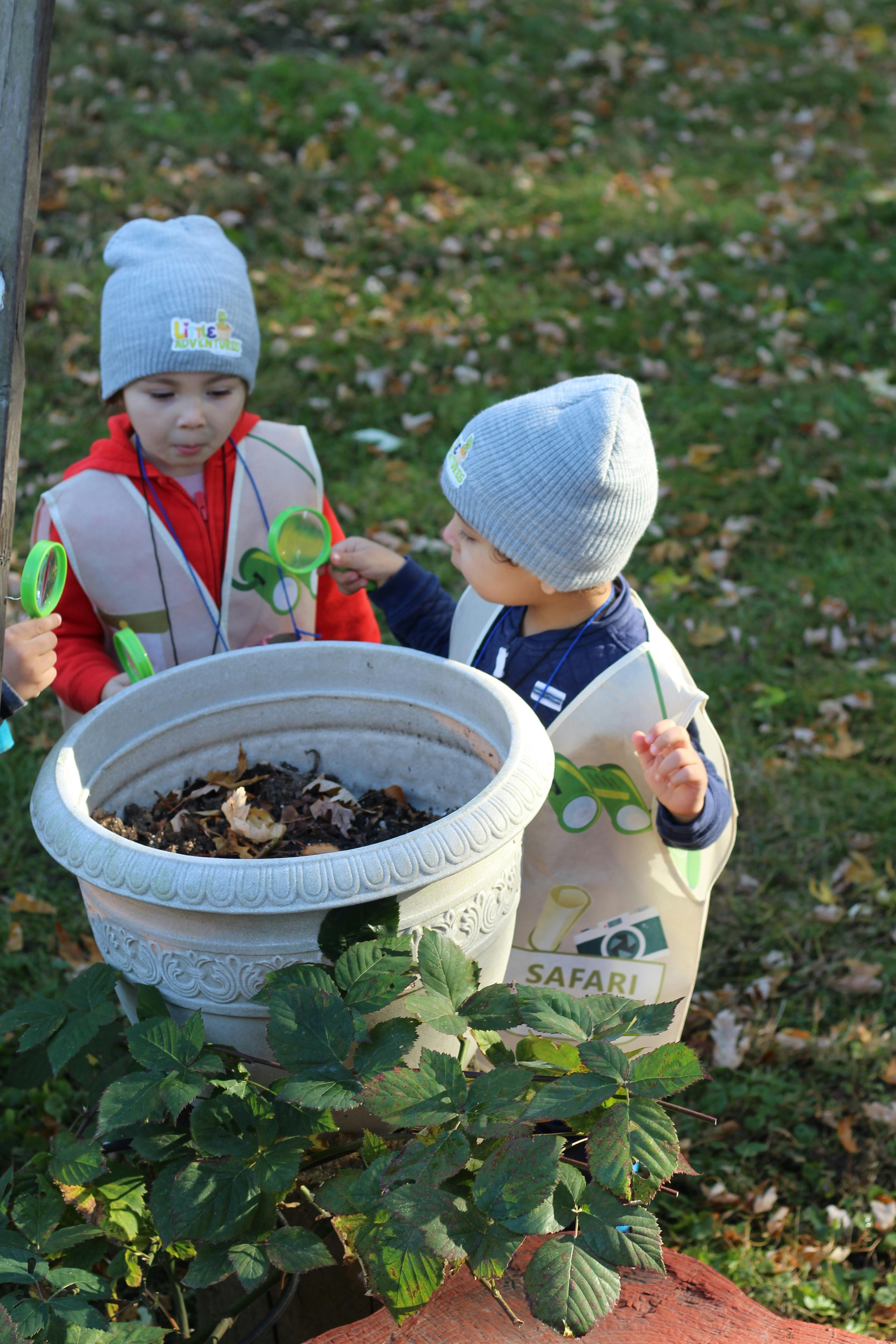 Two children in hats and vests looking into a large planter with magnifying glasses