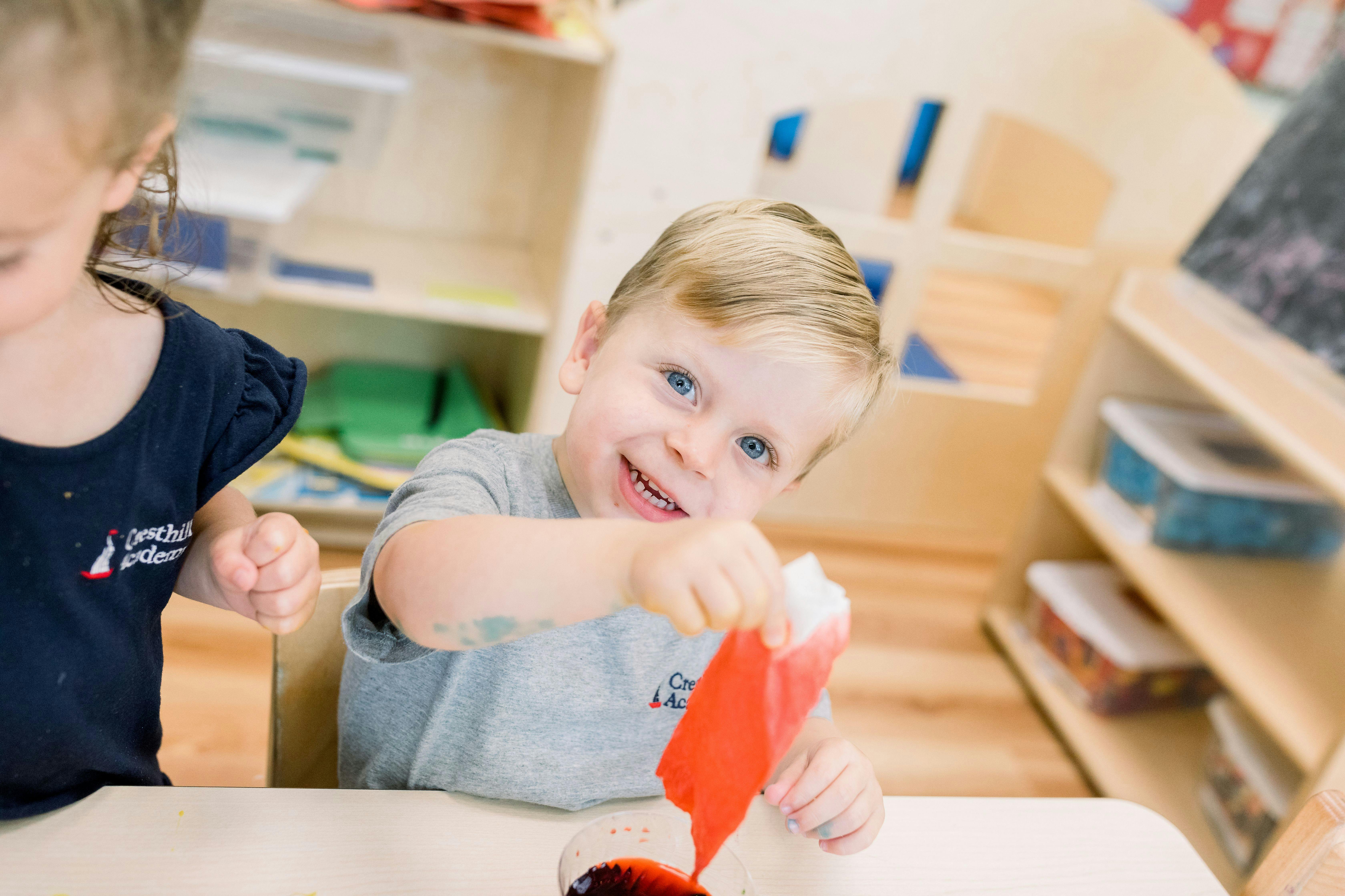Child smiling while holding red paper over a bowl in a classroom