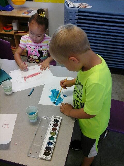 Two children painting and drawing at a table in a classroom