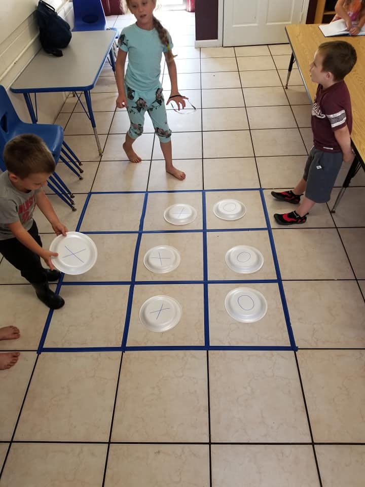 Three children playing tic-tac-toe with plates on a tiled floor