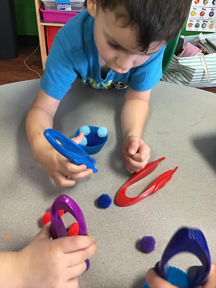 Child using blue tongs to pick up pom-poms from a bowl