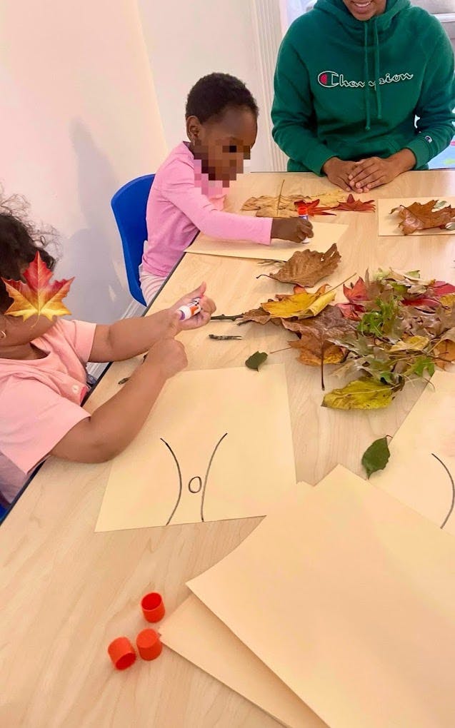 Child gluing leaves onto paper with an adult watching in a classroom