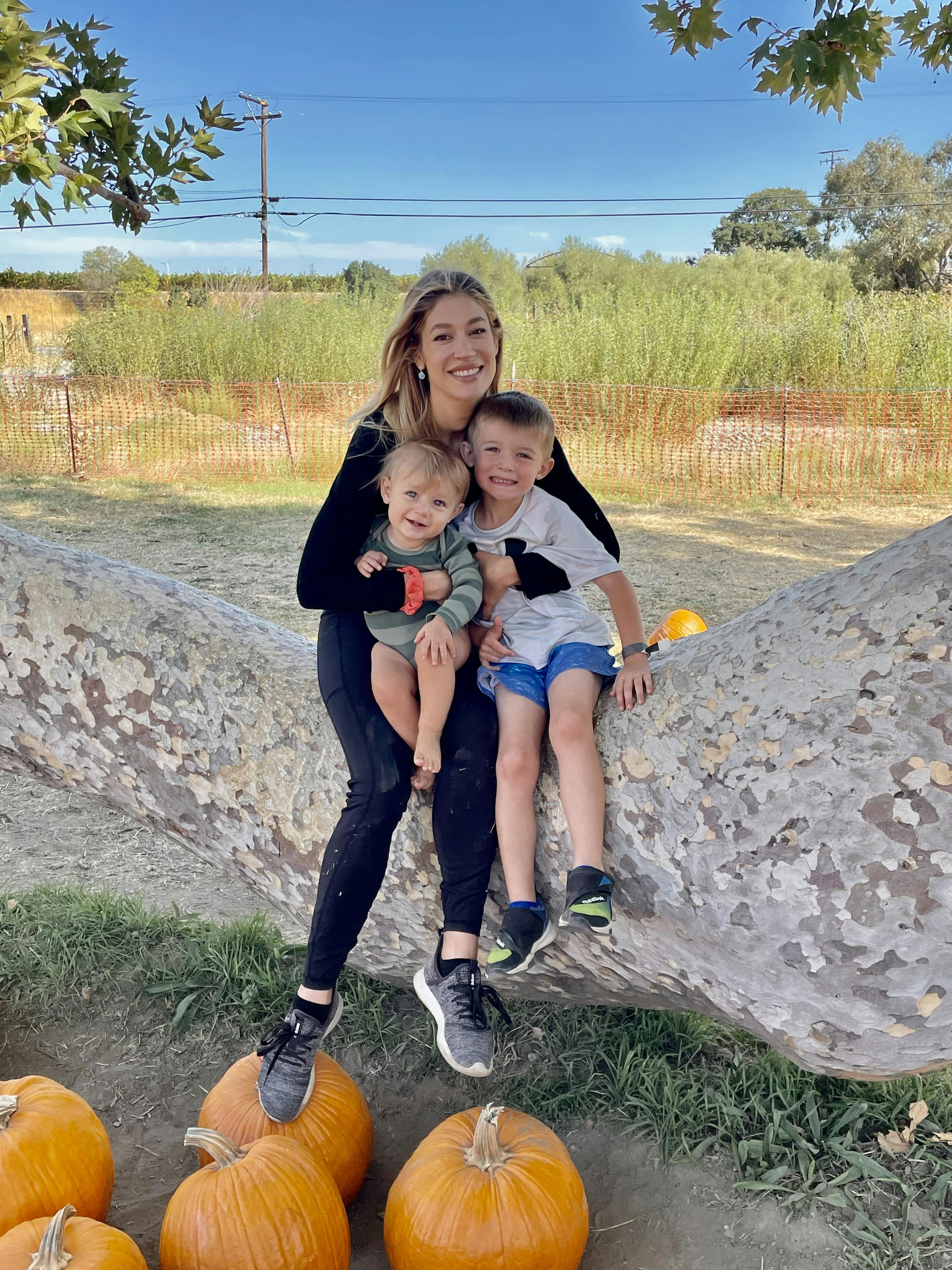 Person holding a child and sitting next to another child on a large log with pumpkins