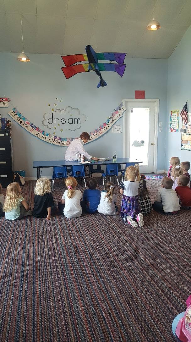 Person at table with children sitting on floor in classroom