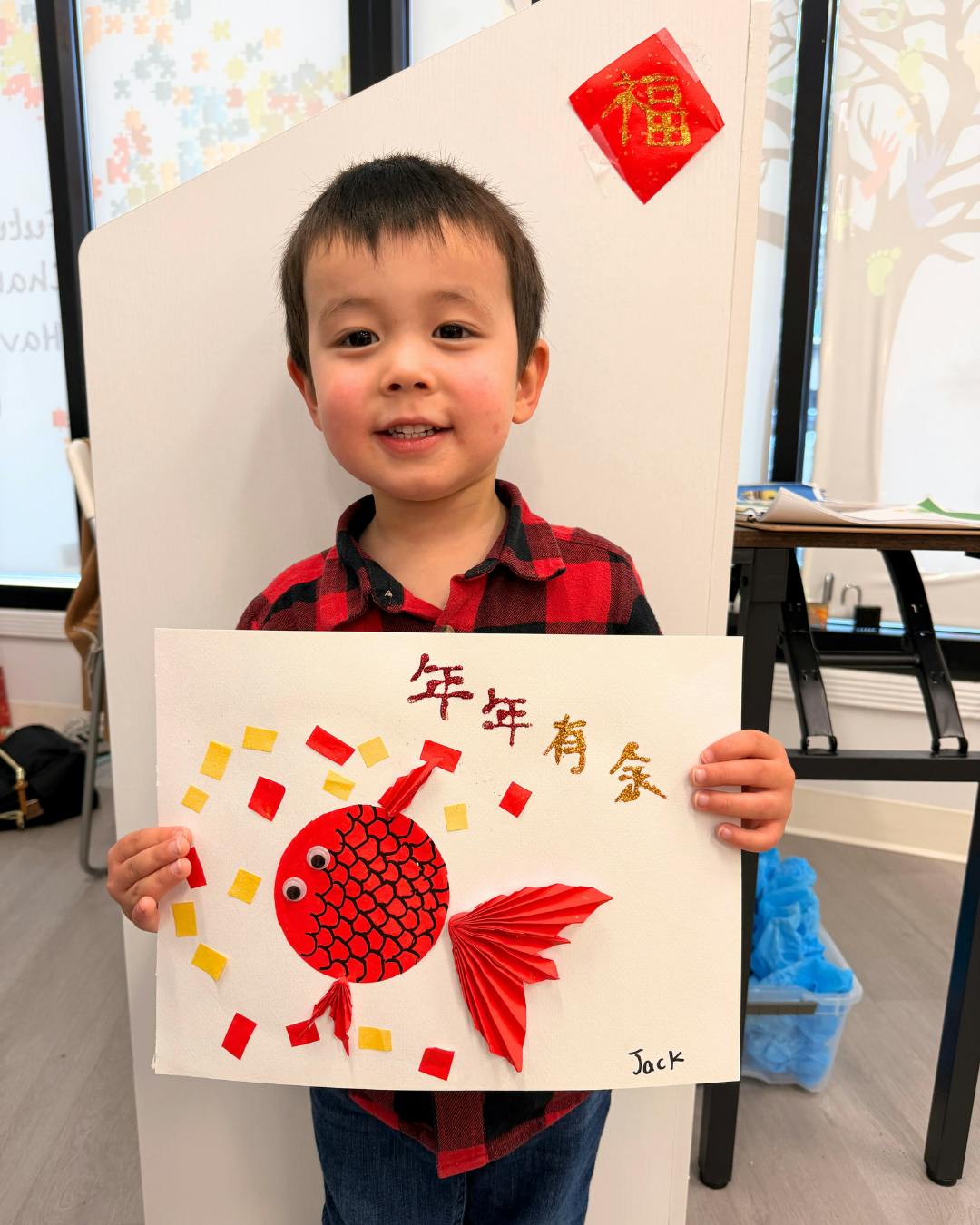 Child in a red plaid shirt holding up a paper craft of a red fish