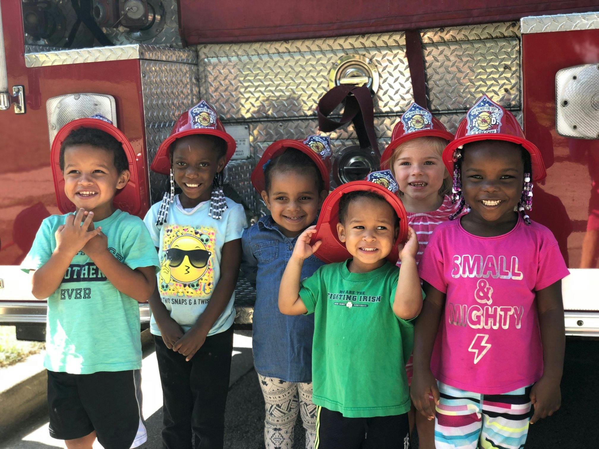 Six children wearing red firefighter hats in front of a fire truck