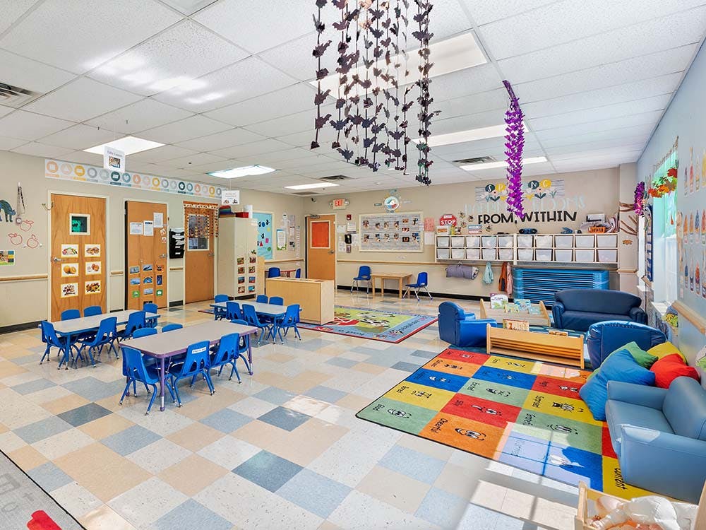 Classroom with small tables and chairs, play area with rugs, and bookshelves