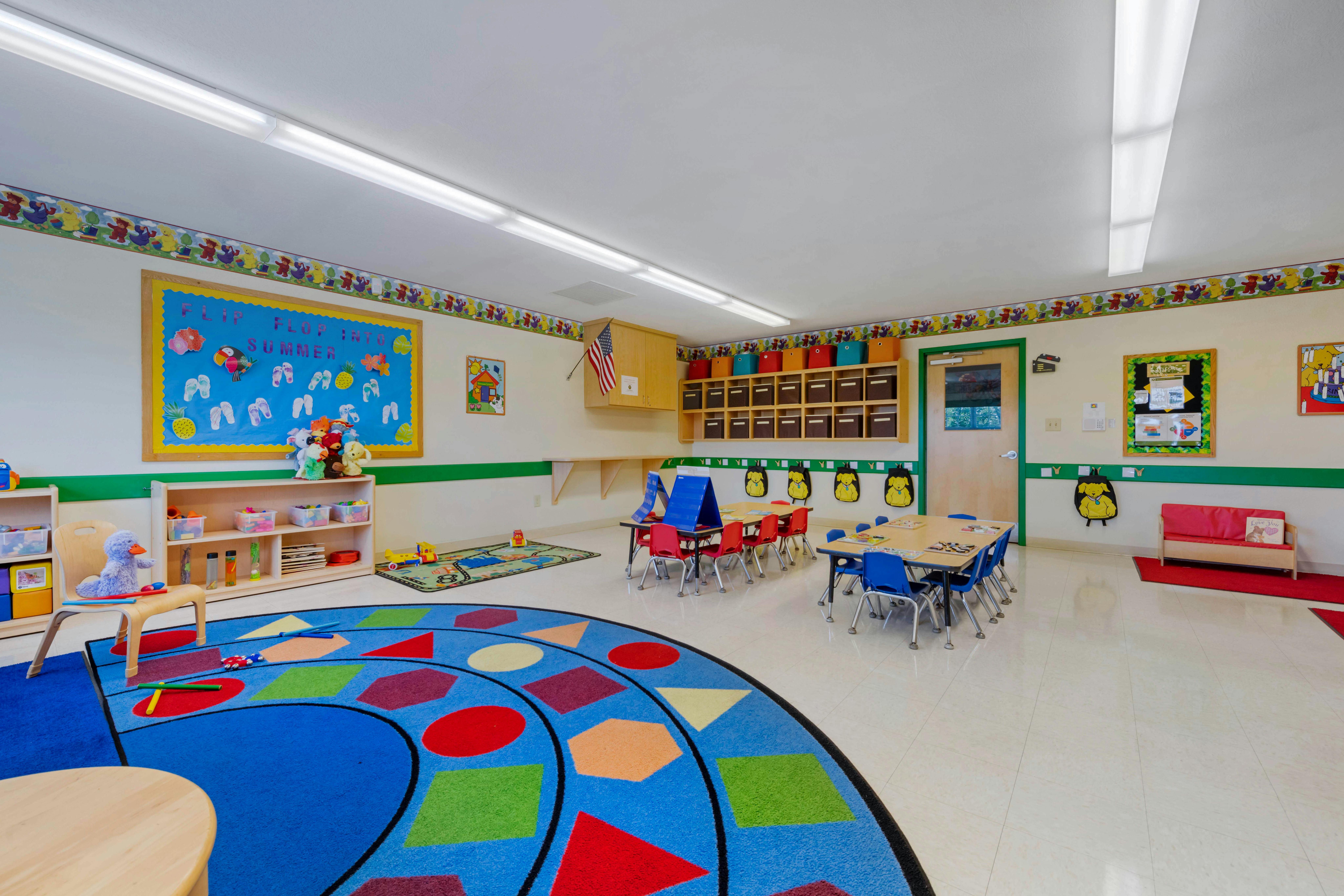 Children's classroom with small tables, chairs, shelves, and a colorful rug