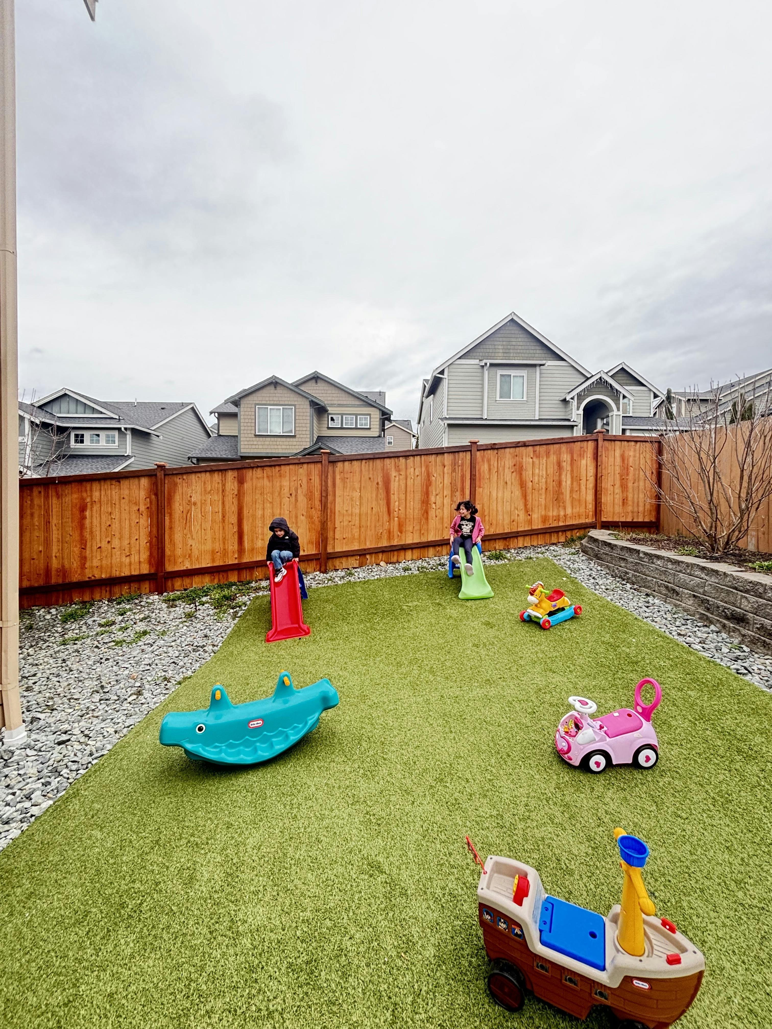 Two children playing on slides in a backyard with artificial turf and toys