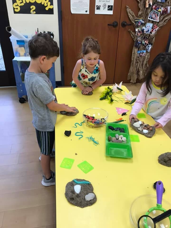 Three children playing with clay and small objects at a yellow table
