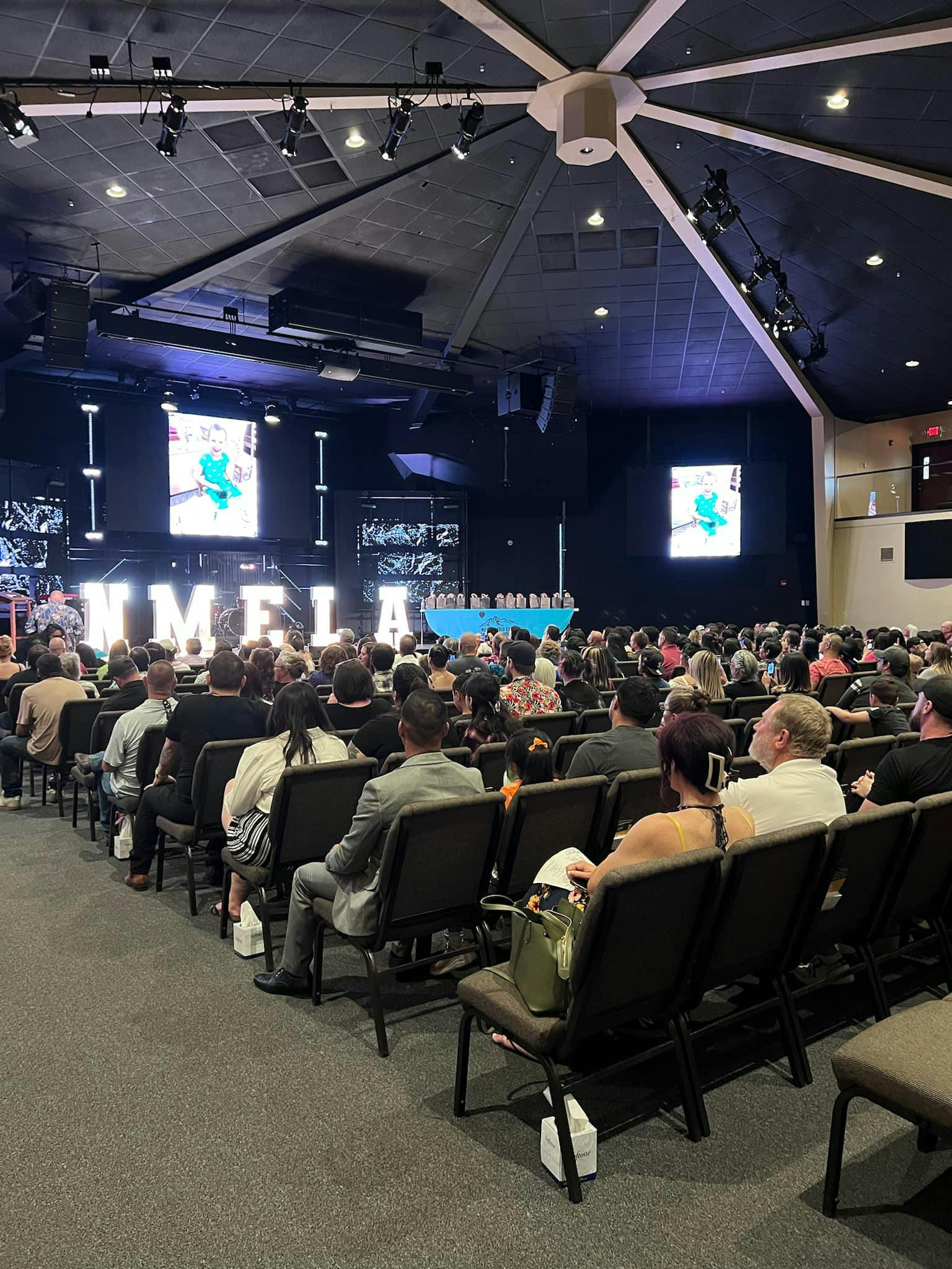 Audience seated in rows facing a stage with large screens and text "WMELA"