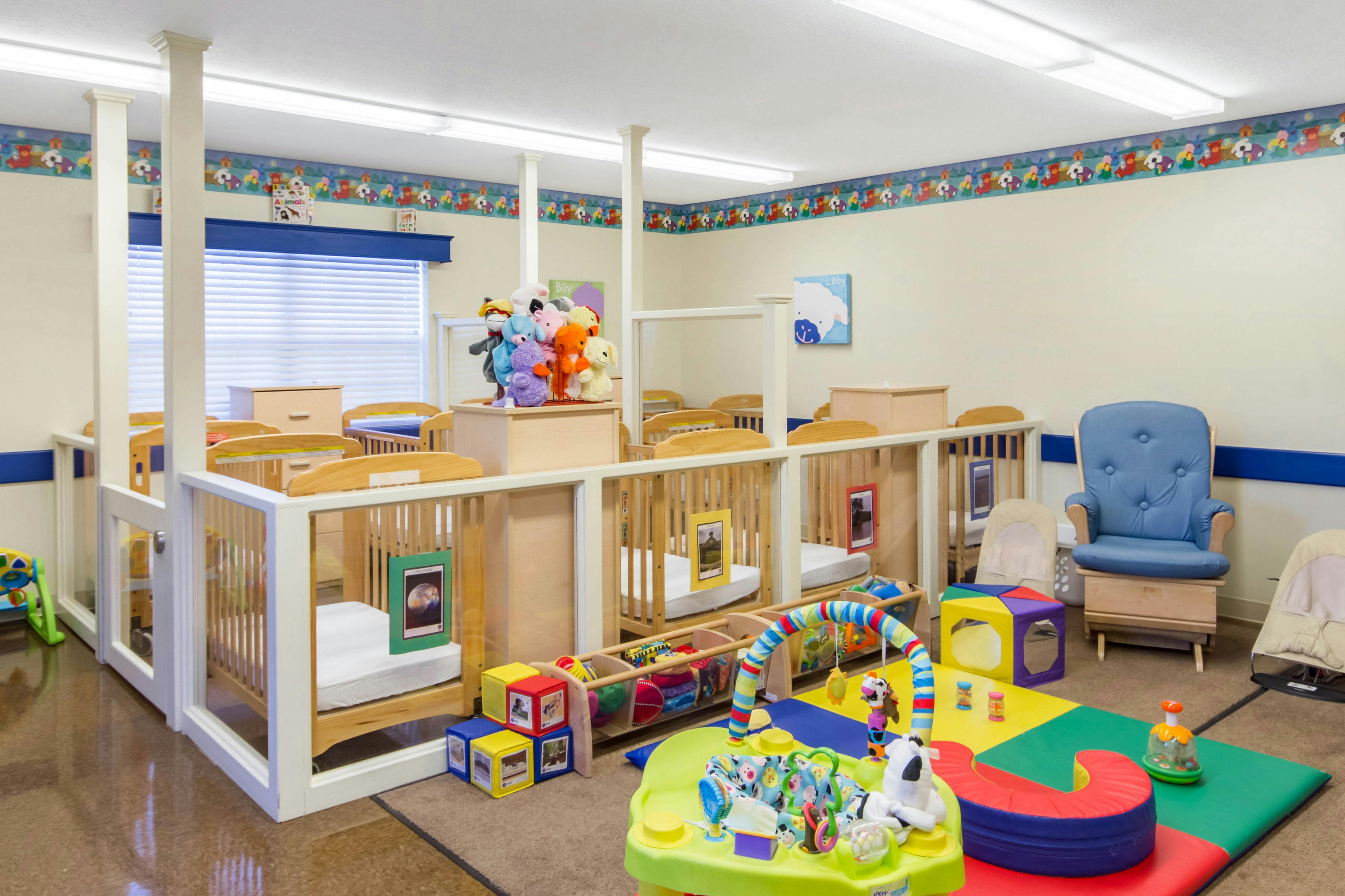 Baby room with multiple cribs, a blue rocking chair, and play mats with toys
