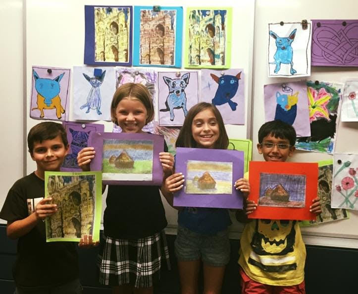 Four children holding up their artwork in front of a wall of other art