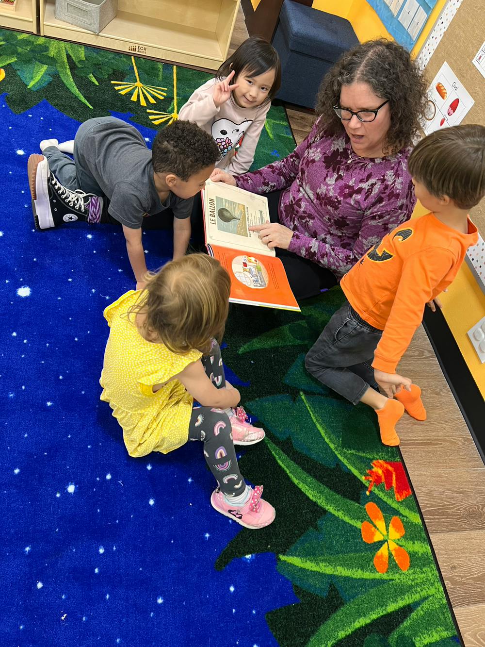 Person reading a book to three children on a colorful rug