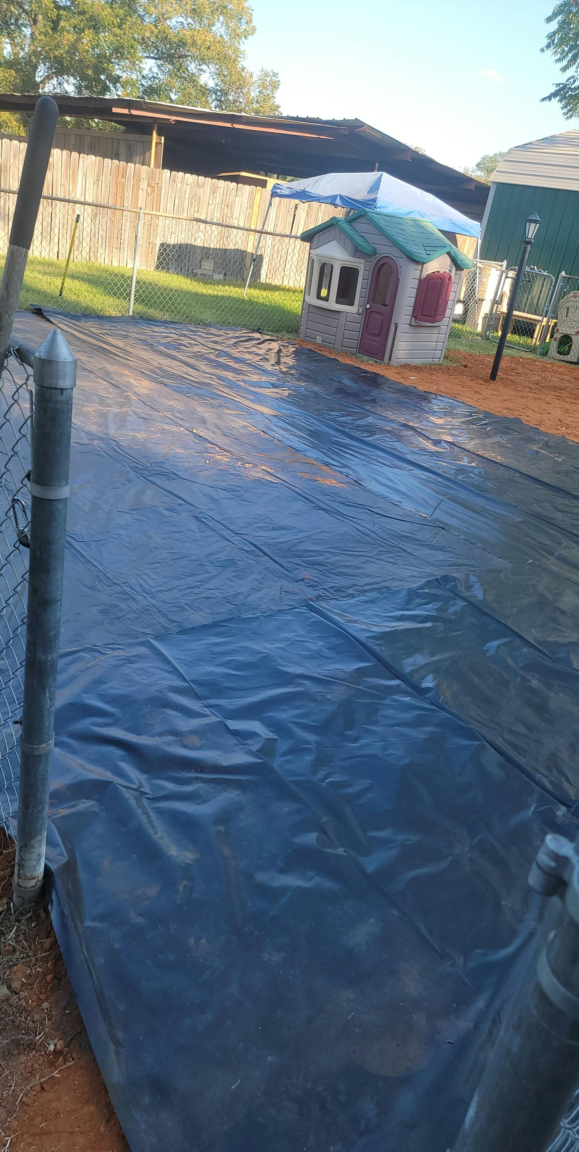 Blue tarp covering the ground next to a chain link fence and a playhouse