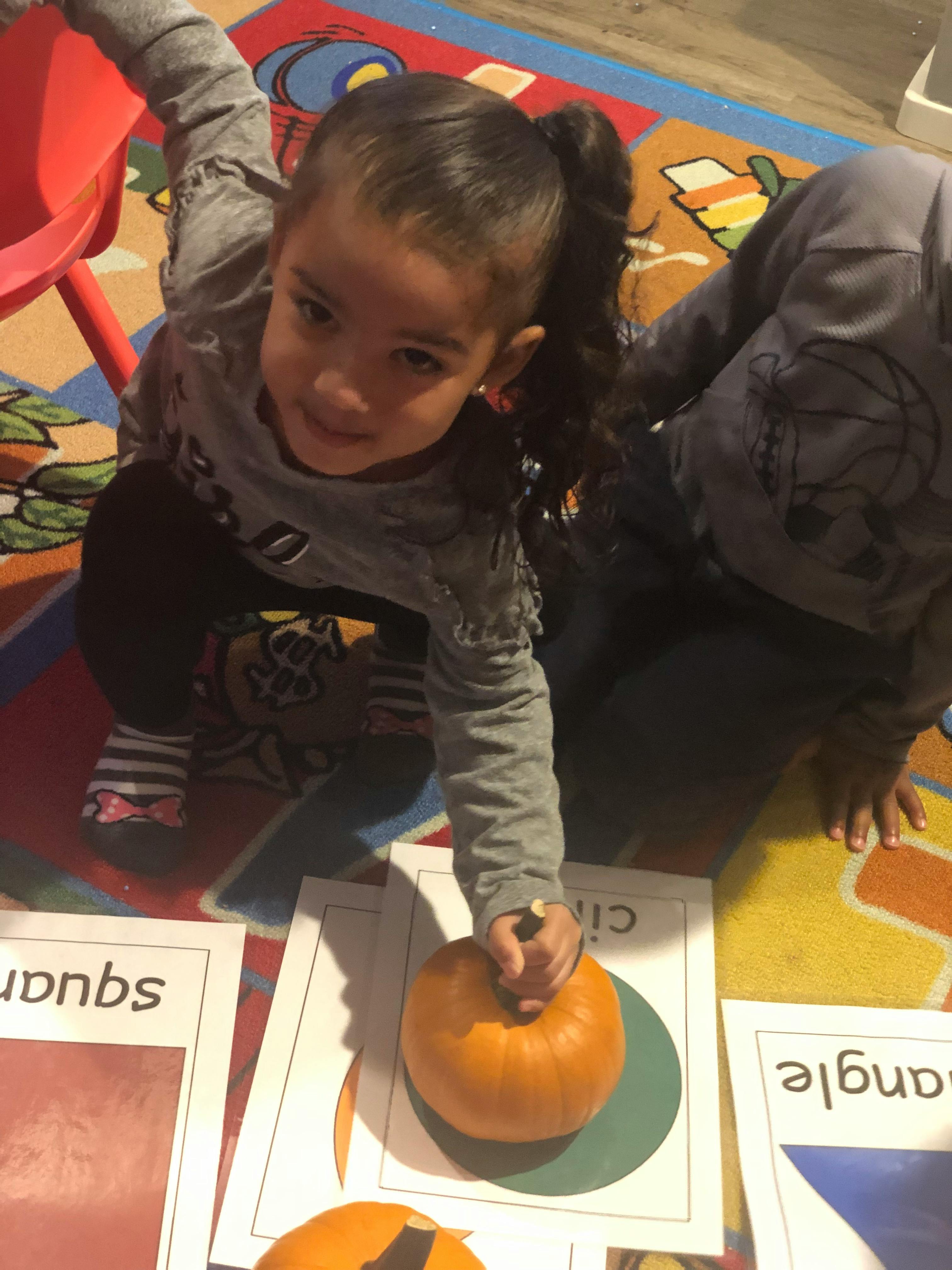 Child holding a small pumpkin over a paper with the word "circle"