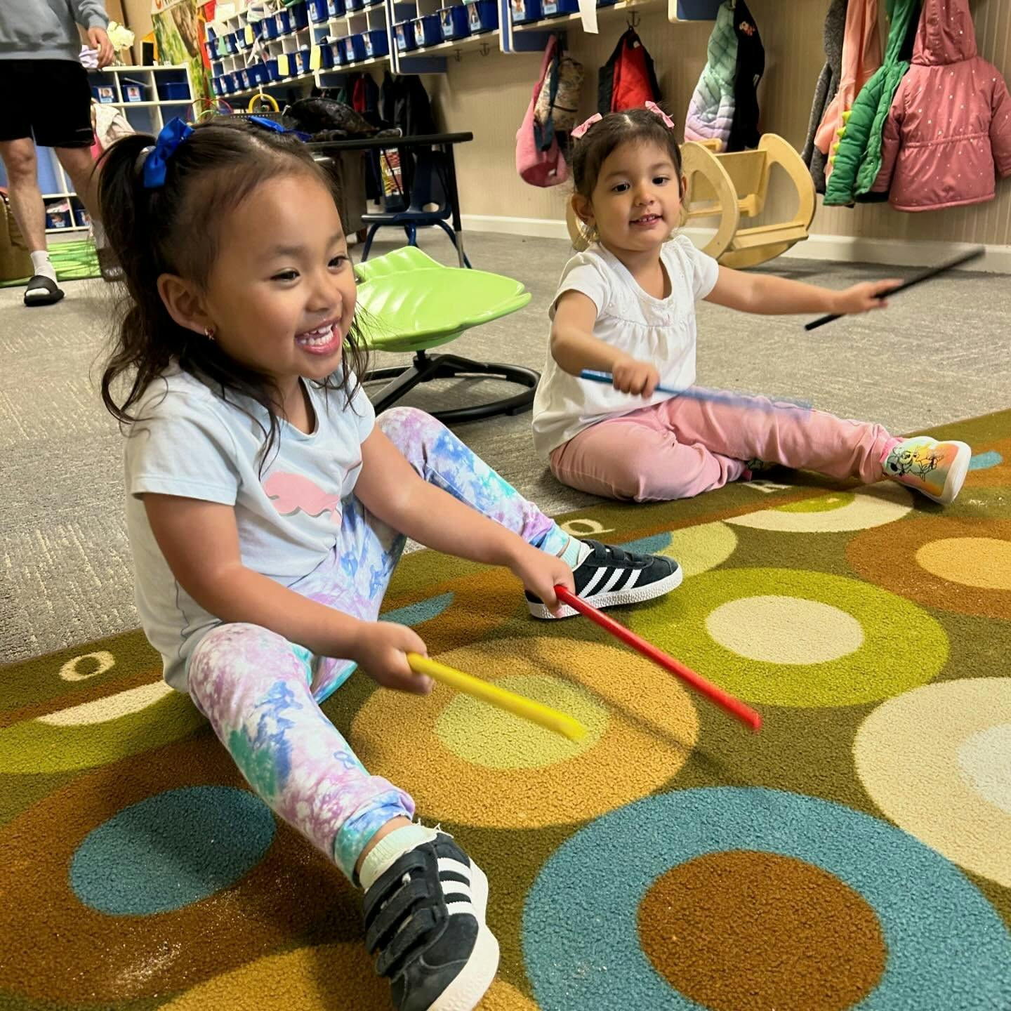 Two children playing with drumsticks on a patterned classroom rug
