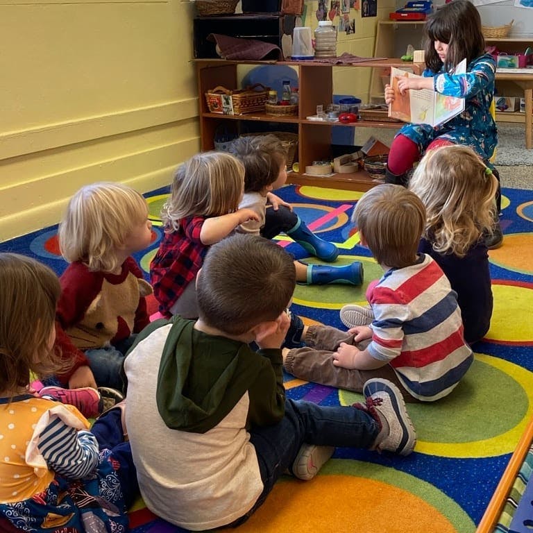 Children sitting on a colorful rug listening to a child read a book