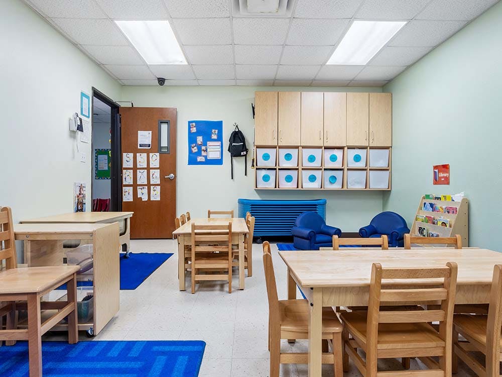 Empty classroom with small wooden tables and chairs, blue rugs, and storage cabinets