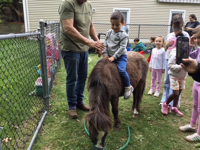 Adult helping a child sit on a small horse with other children nearby