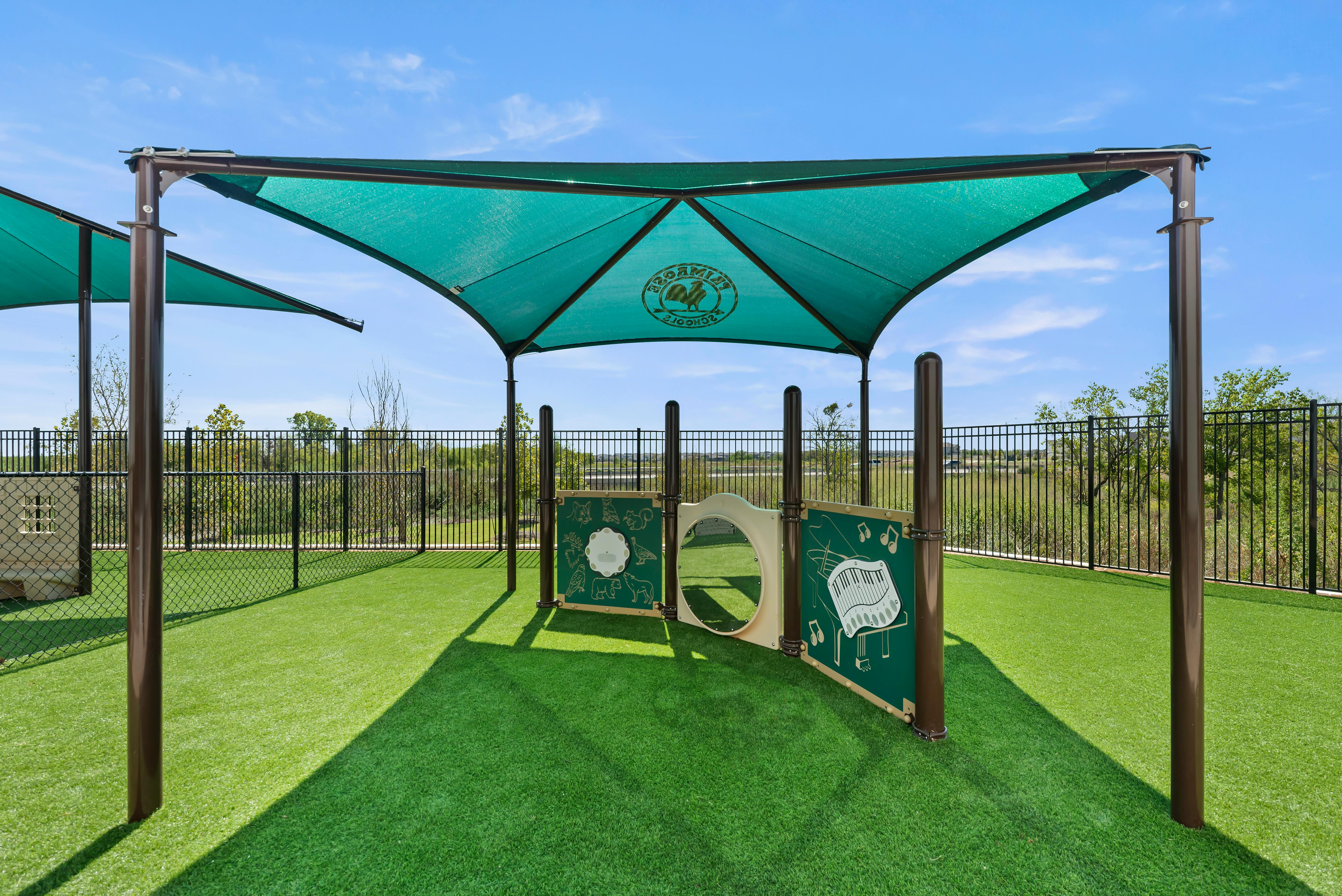 Playground equipment under a green canopy with a fence in the background