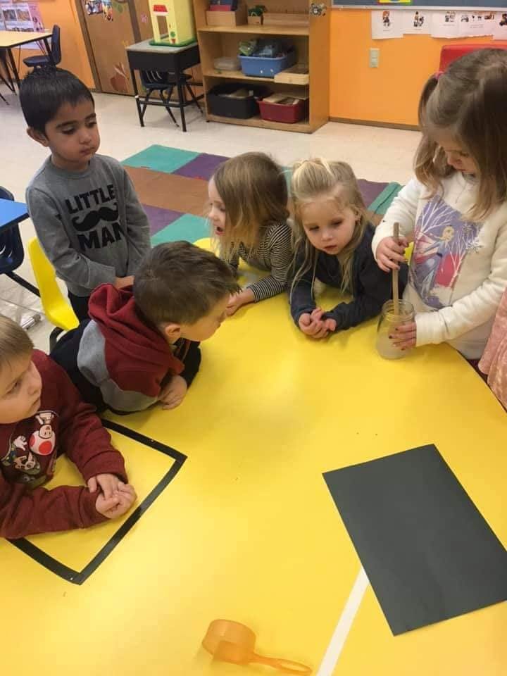 Children gathered around a yellow table, one child stirring a liquid in a jar