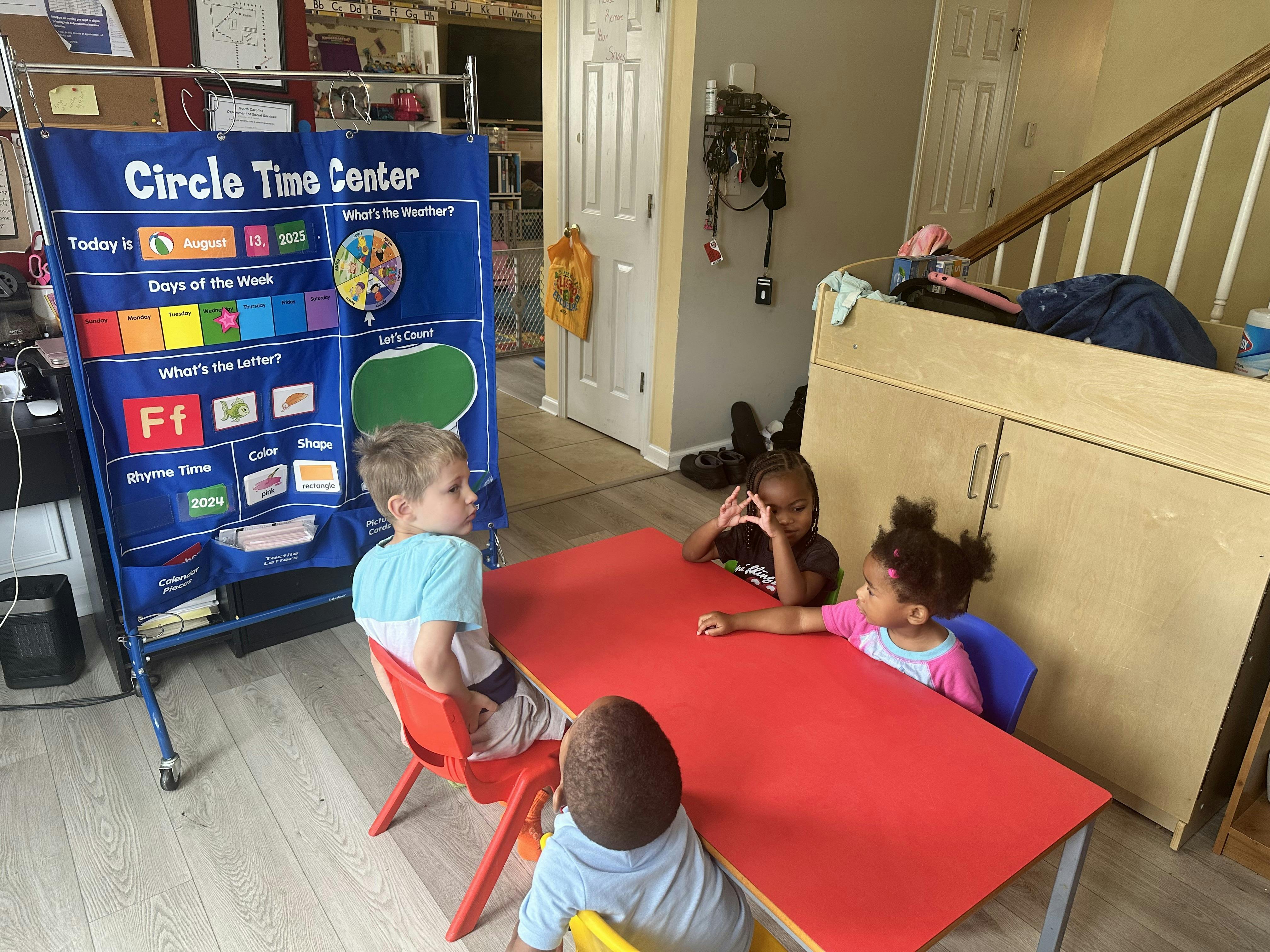 Three children sitting at a red table with a \"Circle Time Center\" calendar in the background