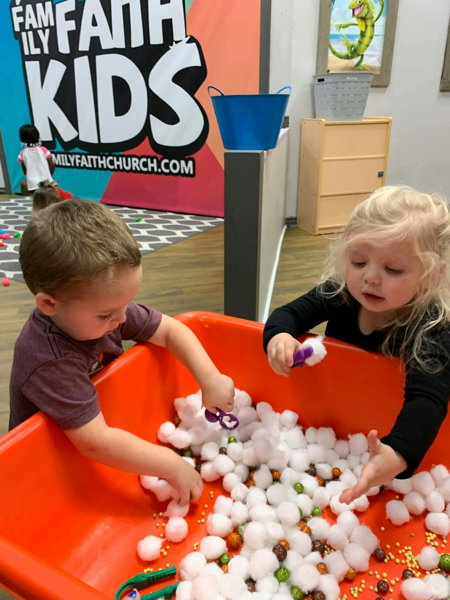 Two children playing with cotton balls and small objects in a bin