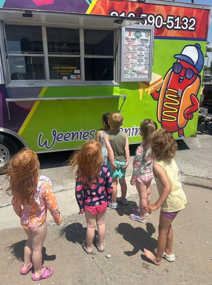 Children waiting in line at a food truck with "Weenies" and a hot dog character