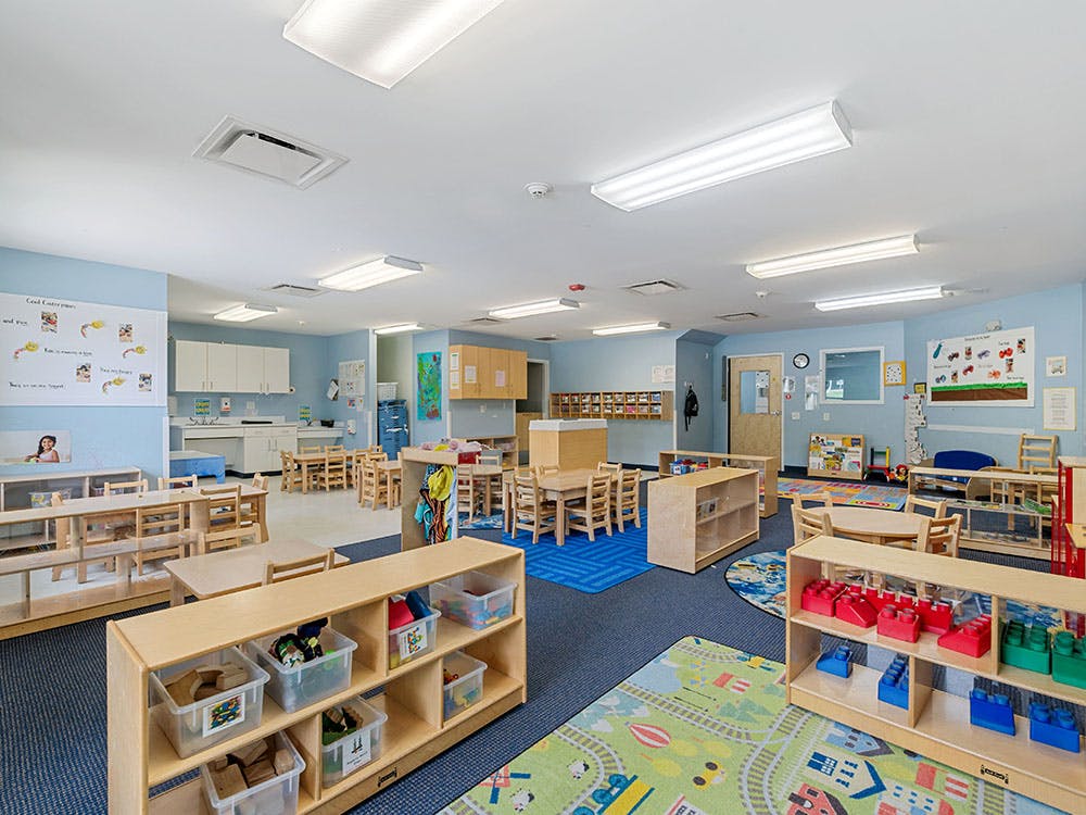 Empty classroom with small wooden tables and chairs, shelves with toys
