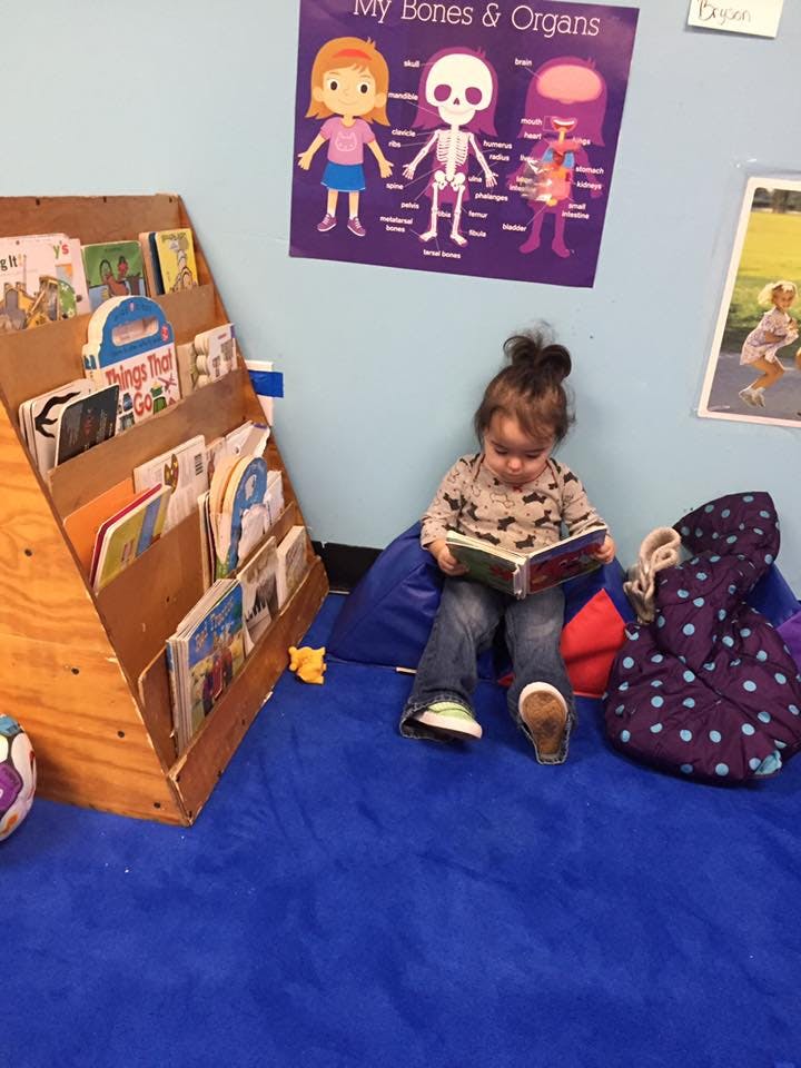 Child sitting on a blue mat reading a book next to a bookshelf