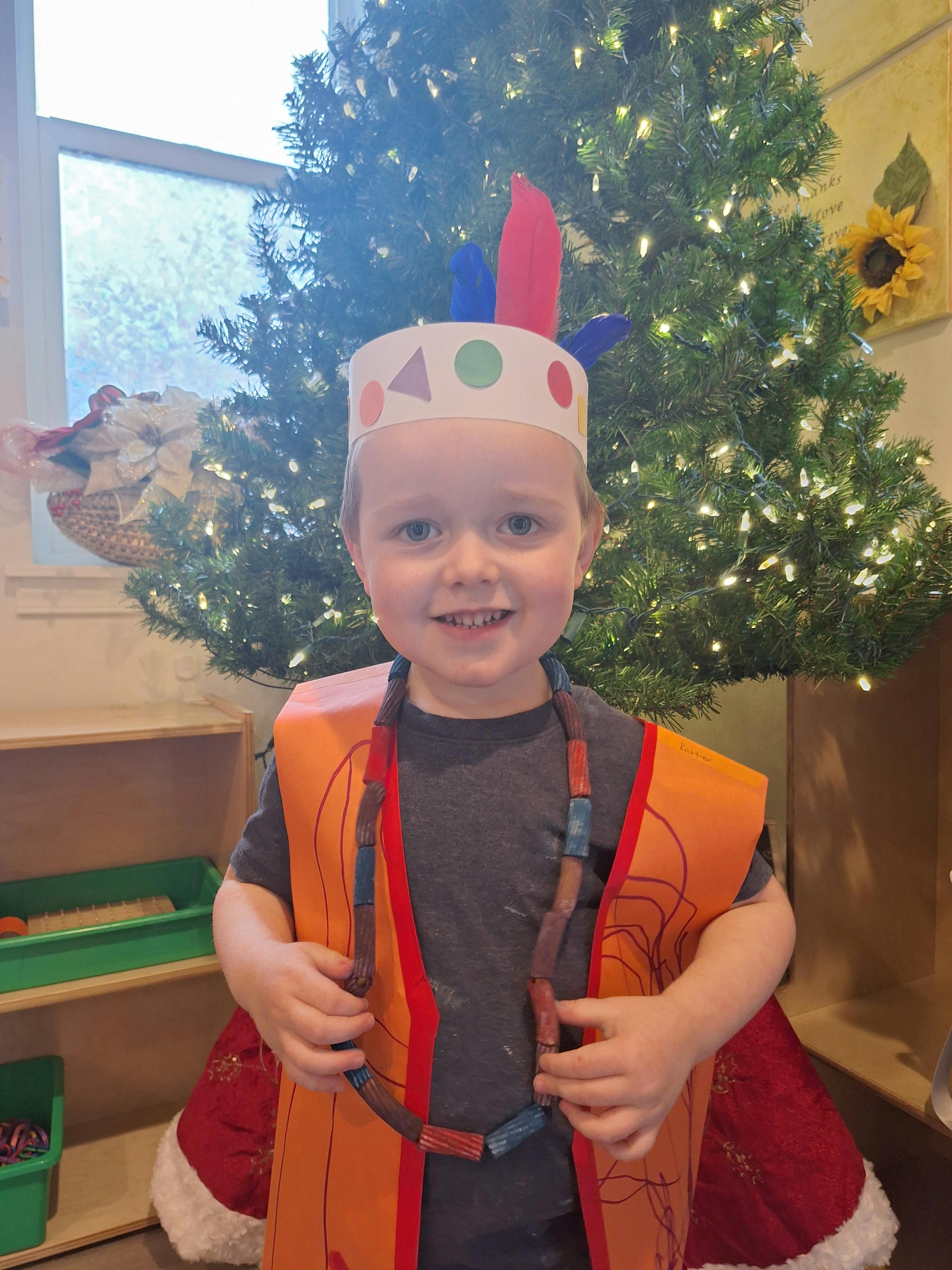 Child smiling in a paper feather headband and orange vest, holding a noodle necklace
