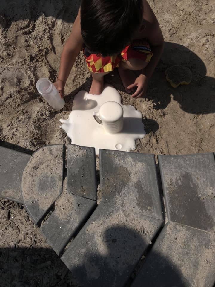 Child pouring liquid from a bottle onto sand next to a cup