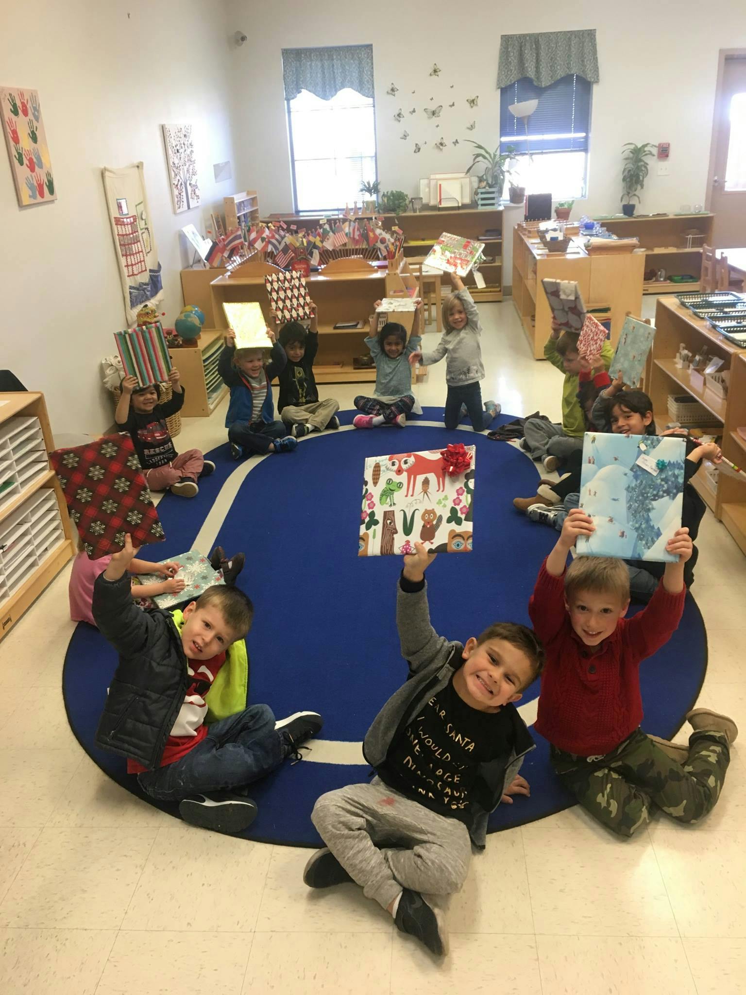 Children sitting on a blue rug holding up wrapped gifts and artwork