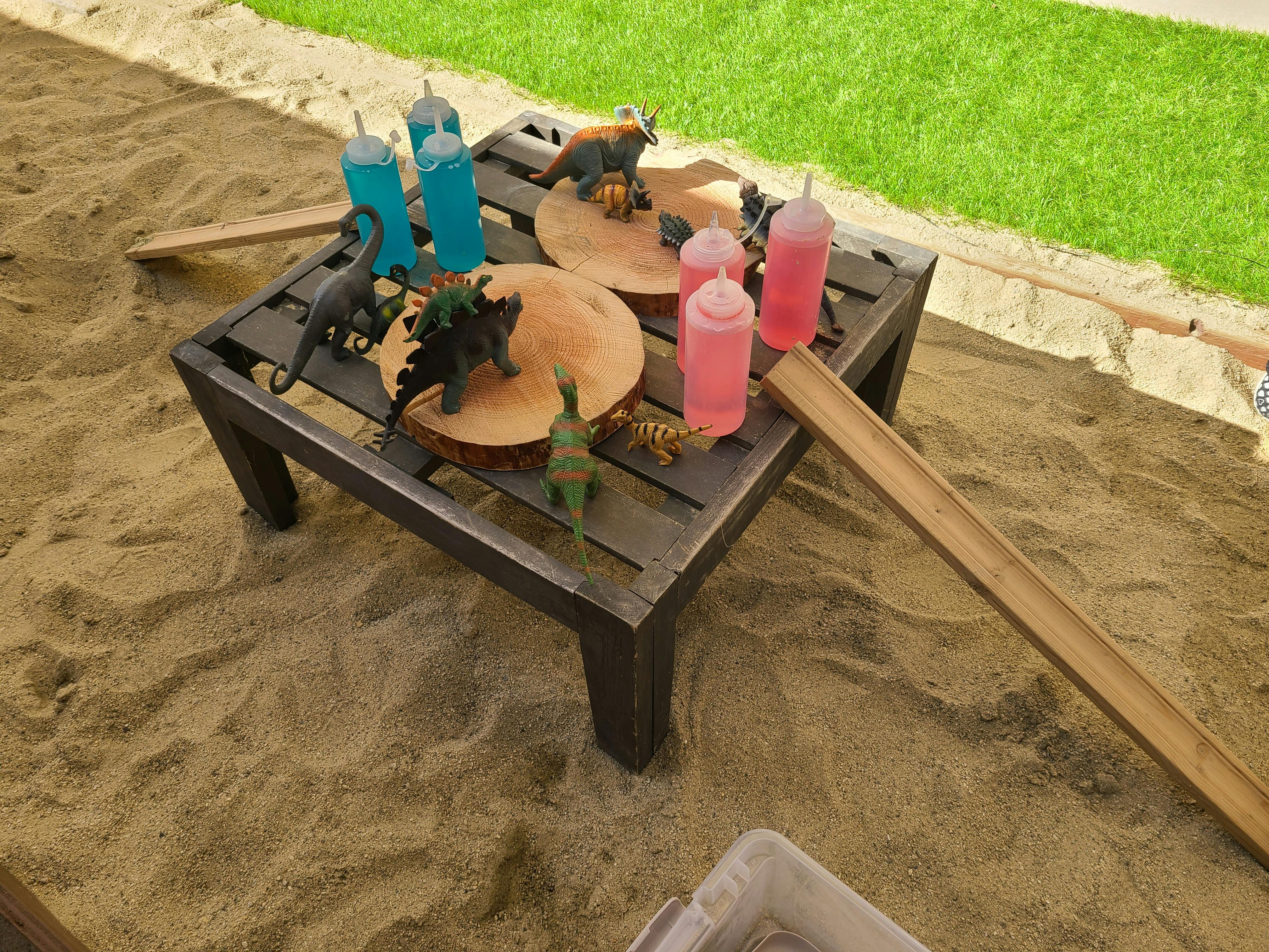 Table with dinosaur toys and bottles of colored liquid in a sandbox