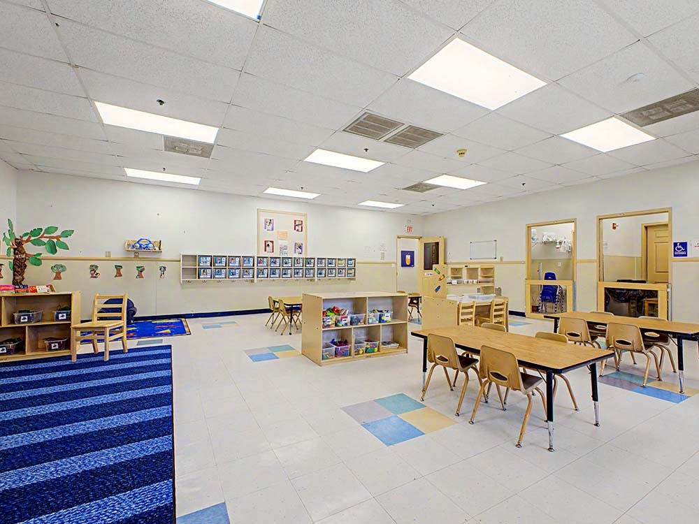 Empty classroom with small tables and chairs, shelves, and a blue striped rug