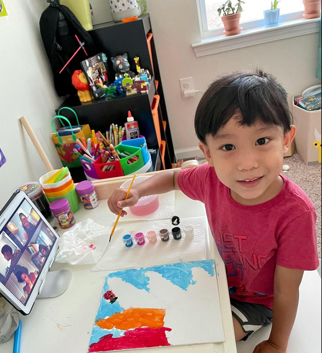 Child painting at a desk with a tablet displaying a video call