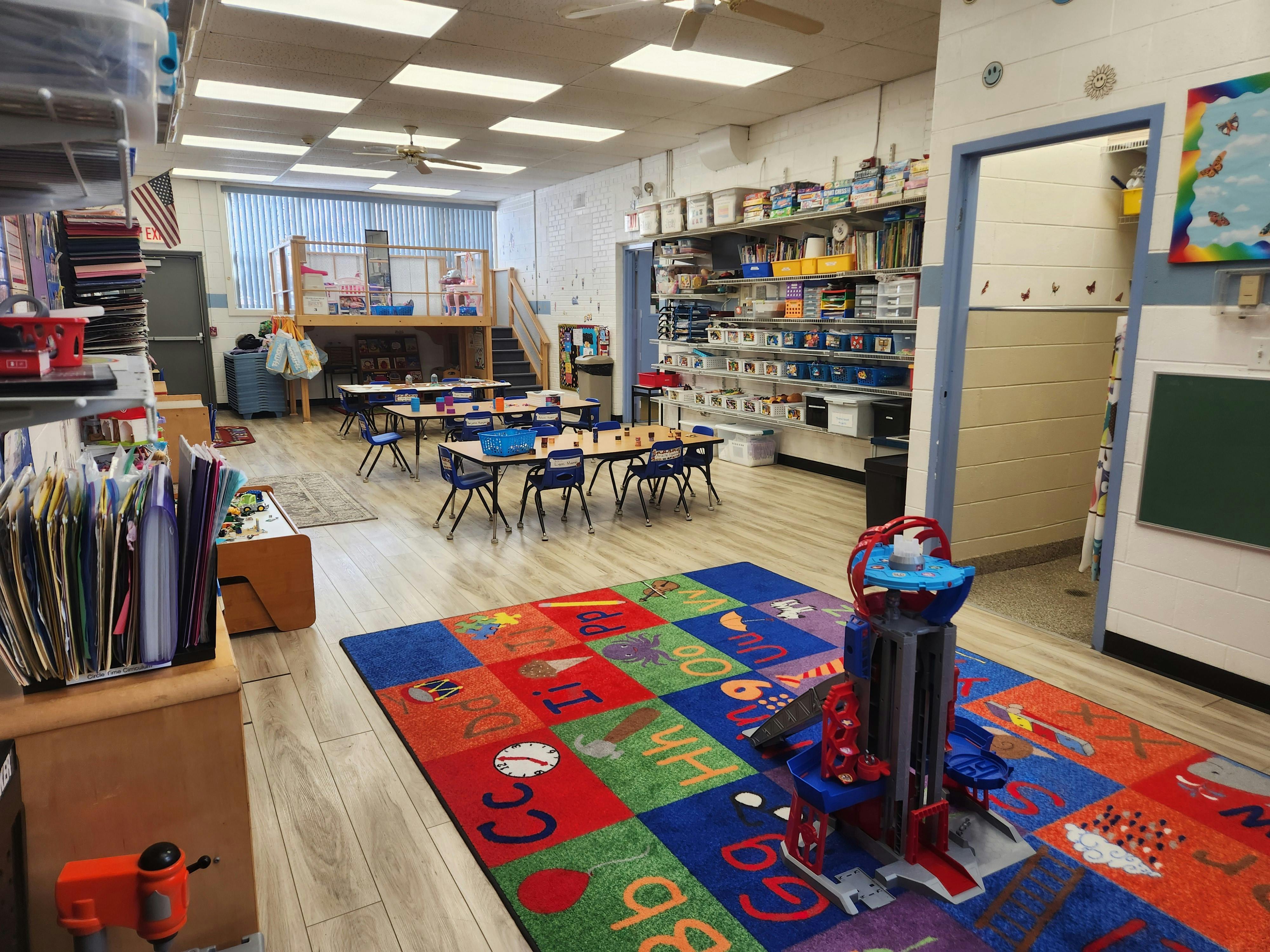 Classroom with small tables and chairs, a colorful rug, and shelves