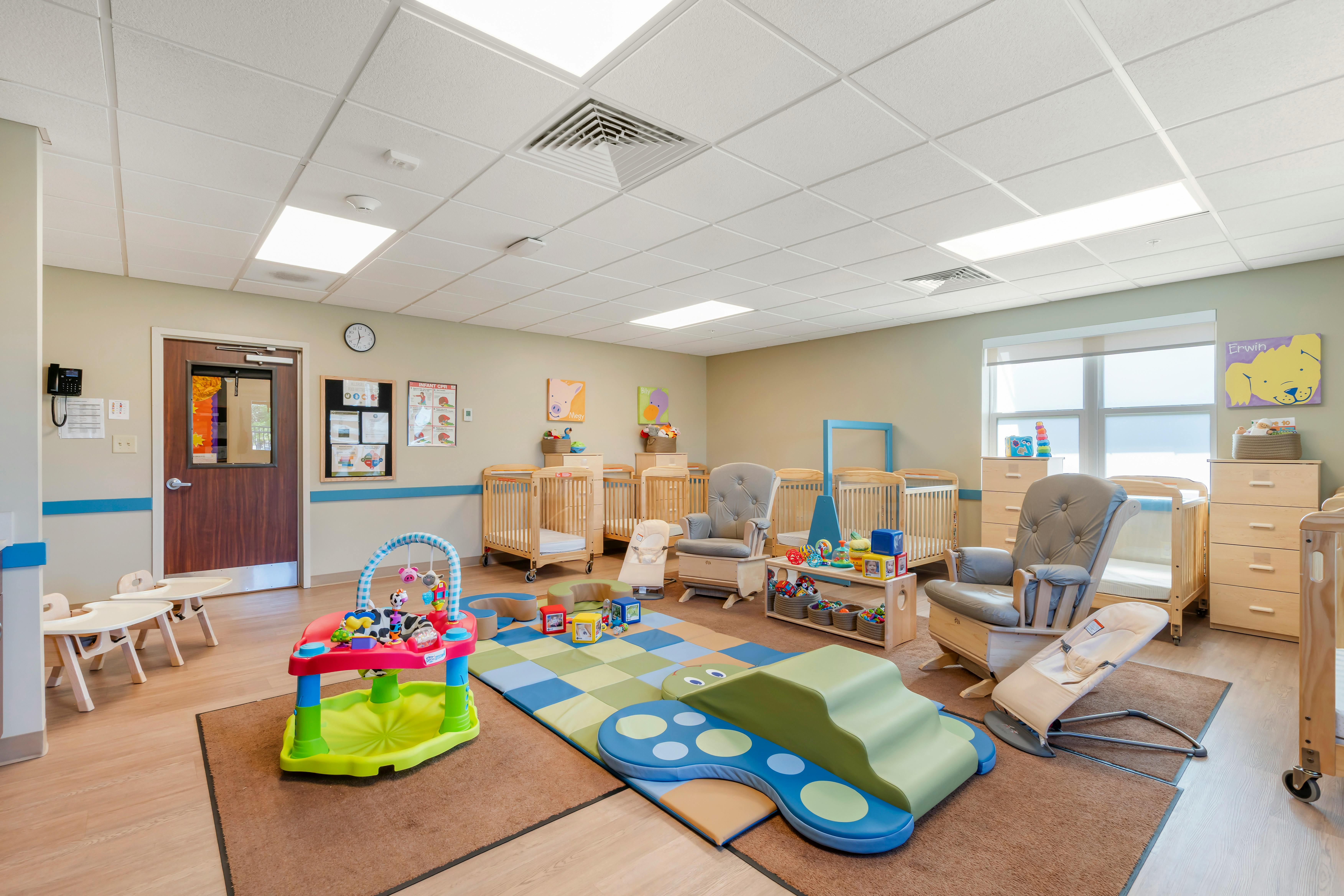 A colorful childcare classroom with cribs, rocking chairs, play mats, and baby toys