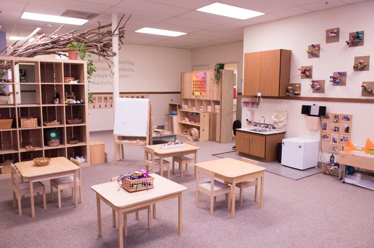 Classroom with small wooden tables and chairs, shelves, and a whiteboard