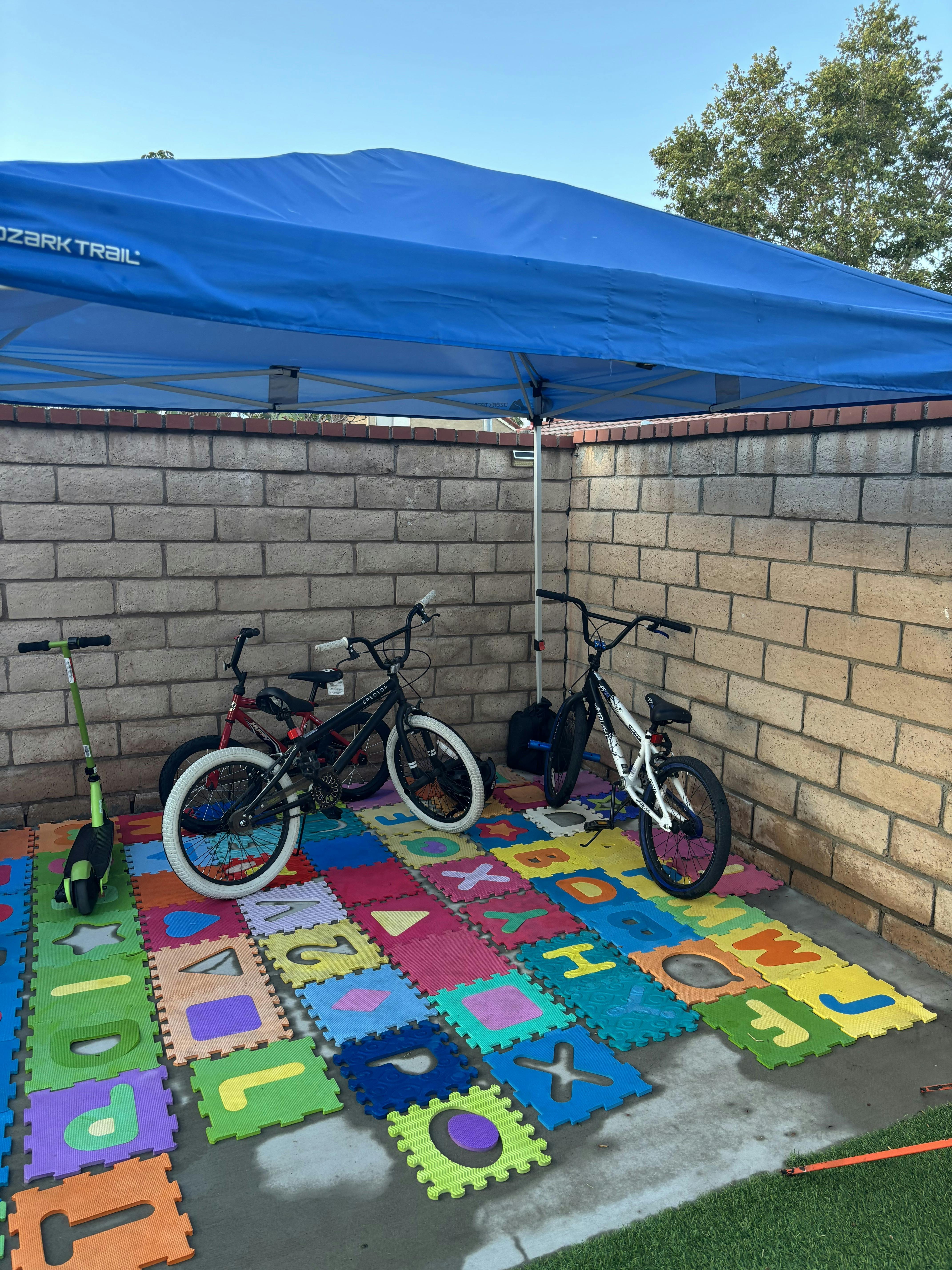 Bicycles and a scooter parked on foam alphabet floor mats under a blue canopy