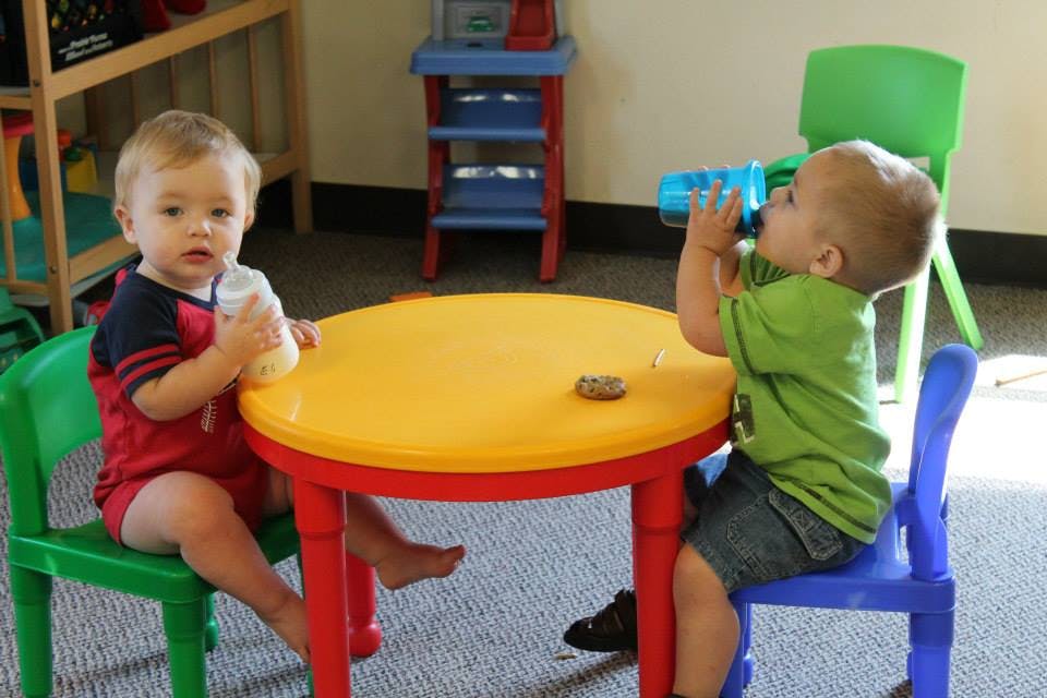Two children sitting at a small table, one drinking from a bottle, one from a sippy cup