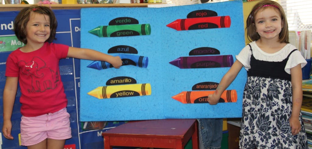 Two children pointing at crayon cutouts on a blue board in a classroom