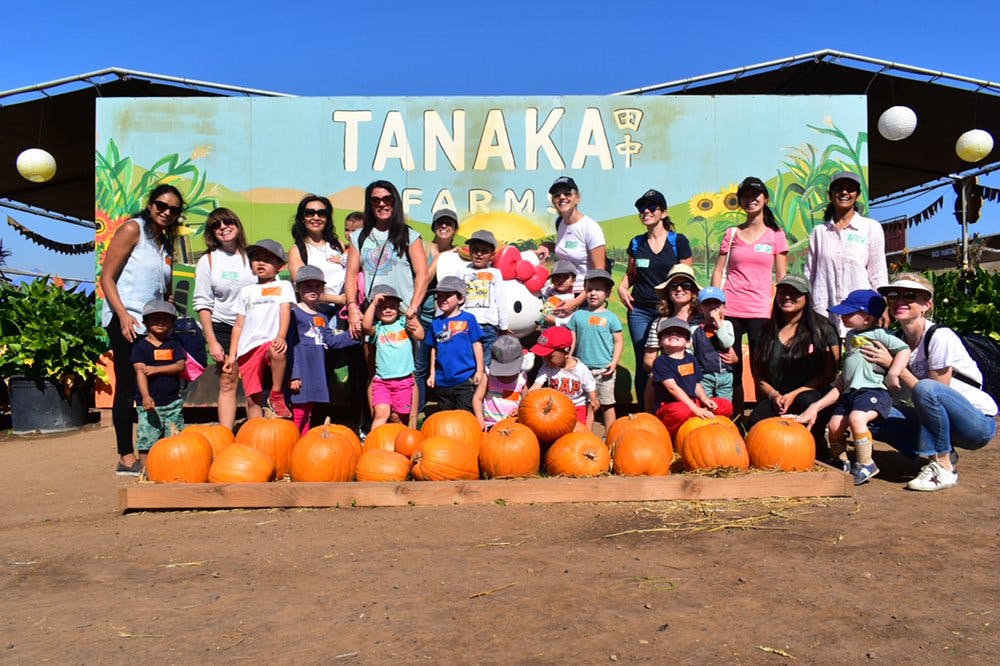 Adults and children posing with pumpkins in front of a "Tanaka Farms" sign