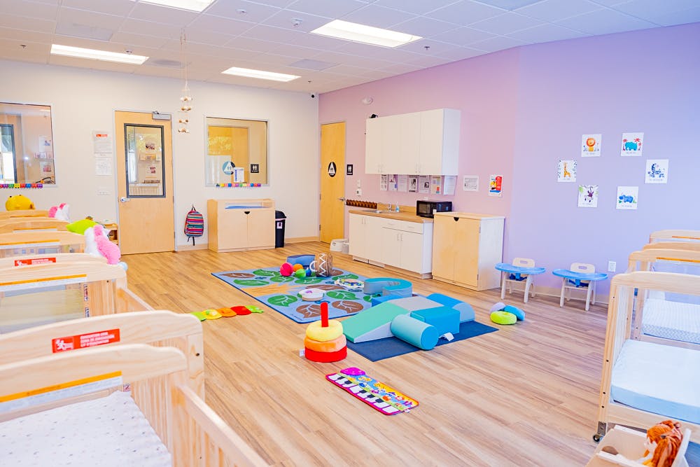 Empty daycare room with cribs, playmat, and small tables