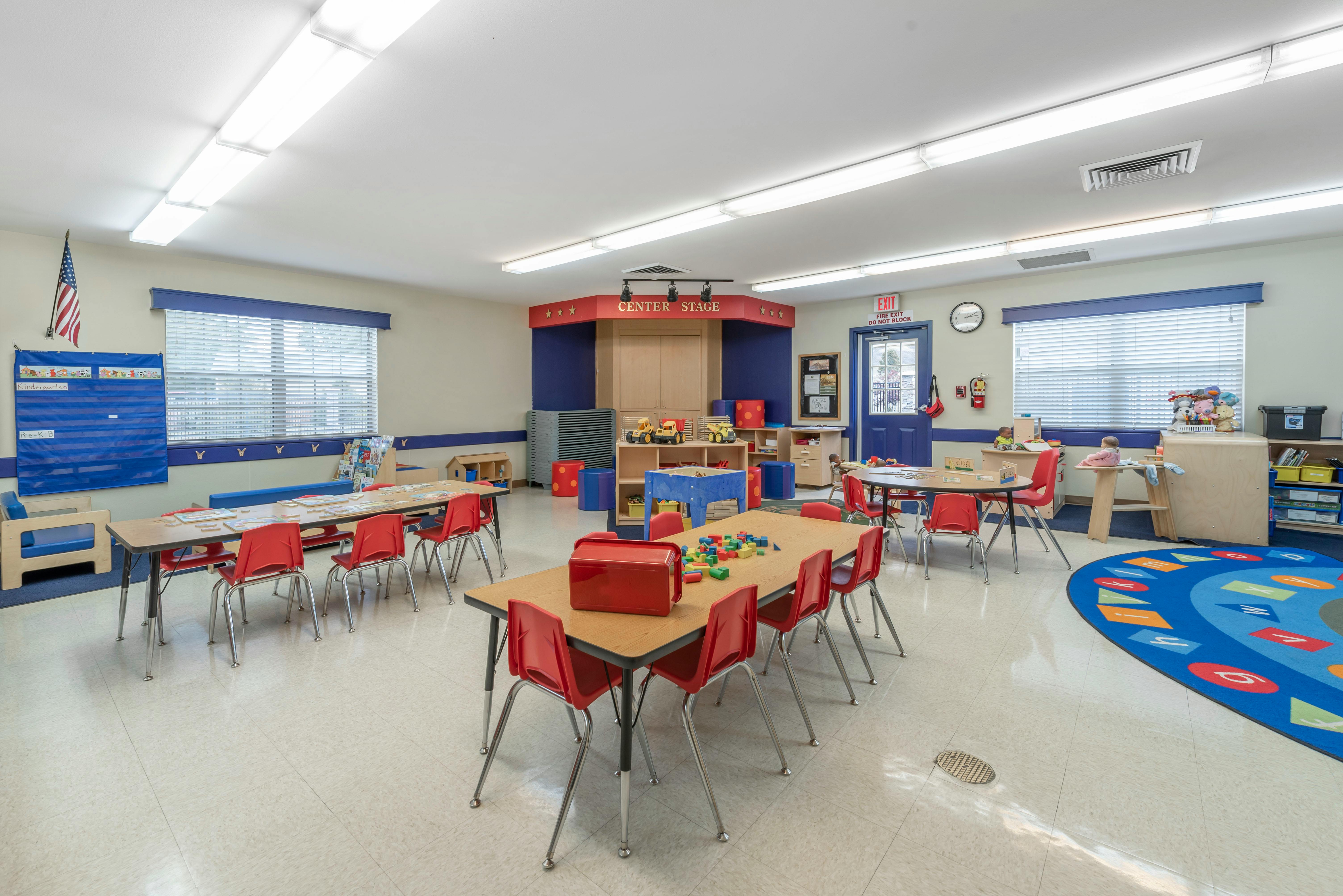 A colorful kindergarten classroom with small tables, red chairs, and play areas