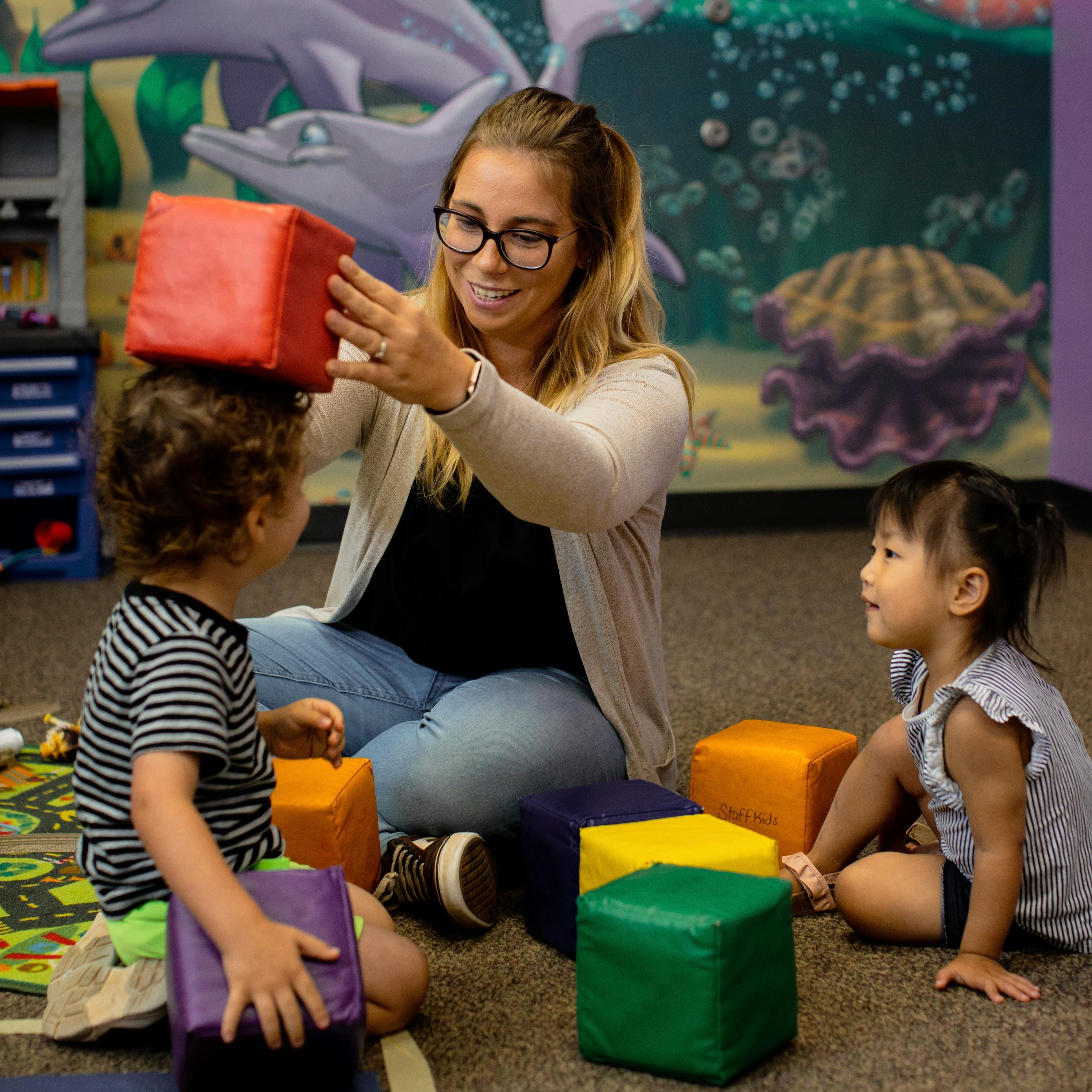 Person placing a red block on a child's head, another child watching